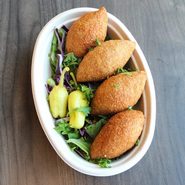Four golden-brown kibbeh served in a white bowl with green lettuce, red cabbage, and yellow peppers.